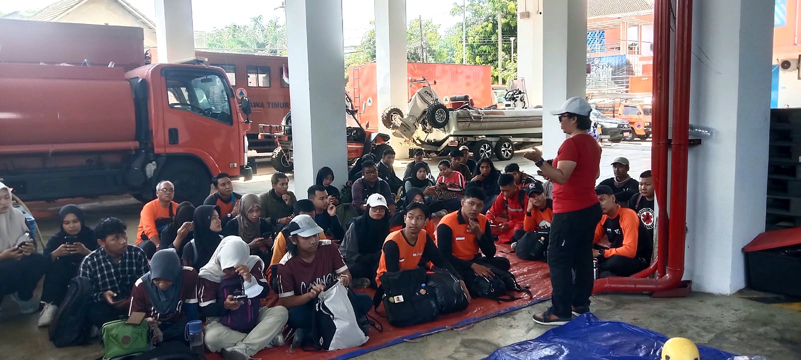 Puluhan peserta berjaket dan berhijab duduk melingkar di lantai mengikuti pengarahan, dengan truk tangki dan peralatan di belakang mereka