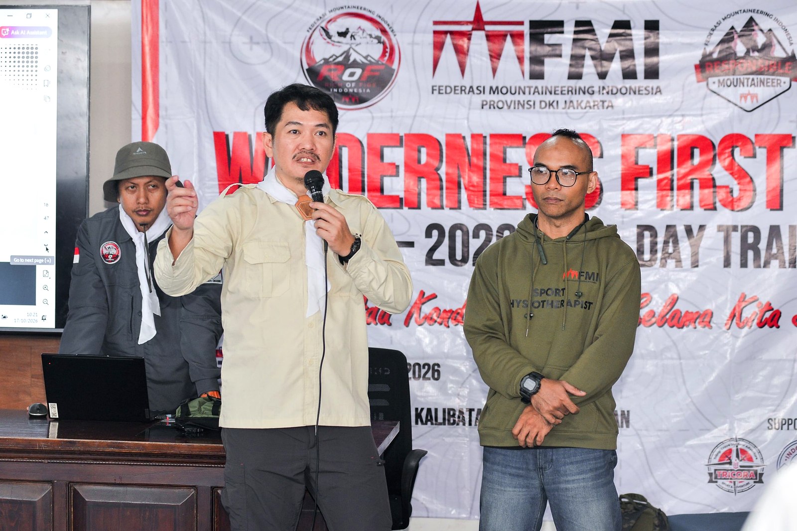 Seorang pria berbicara dengan mikrofon di depan spanduk Wilderness First Aid FMI DKI Jakarta, dua orang berdiri di sampingnya